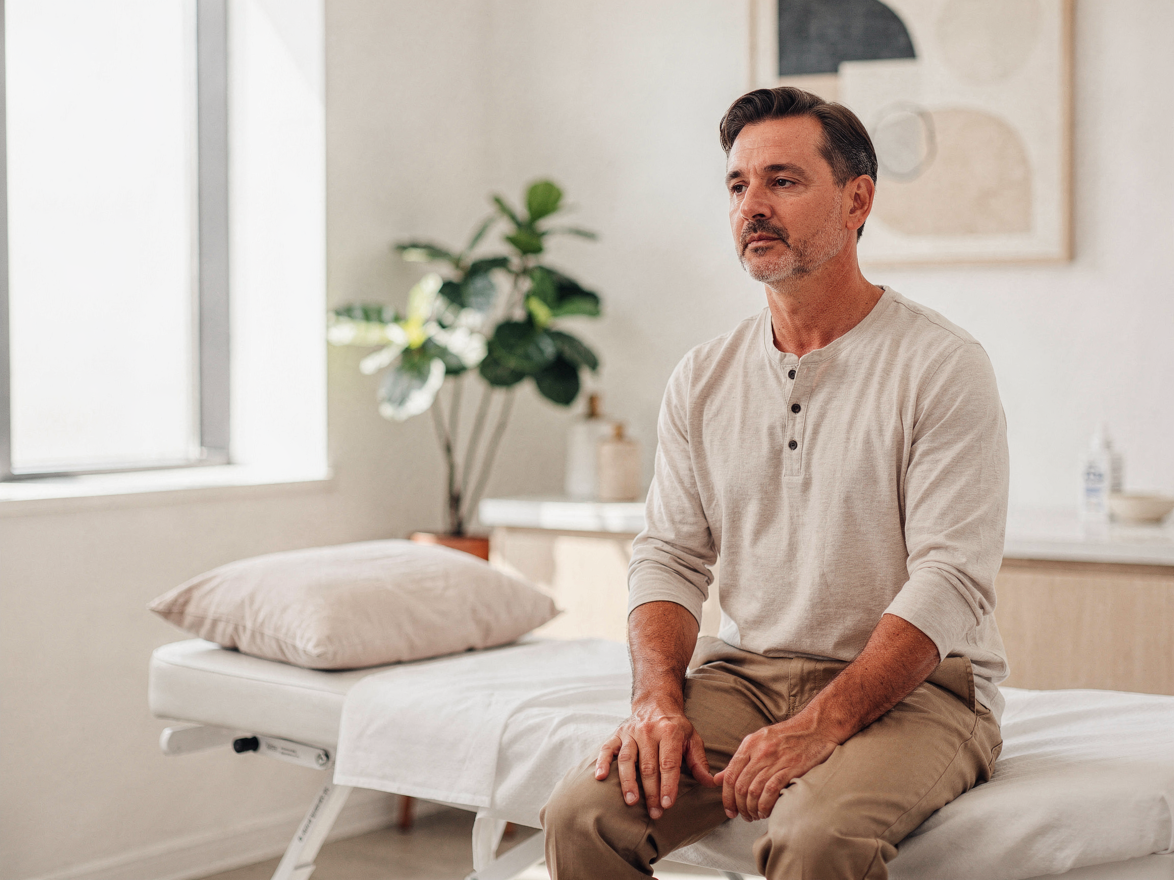 Man sitting on an examination bed in a bright clinical room with plants and windows
